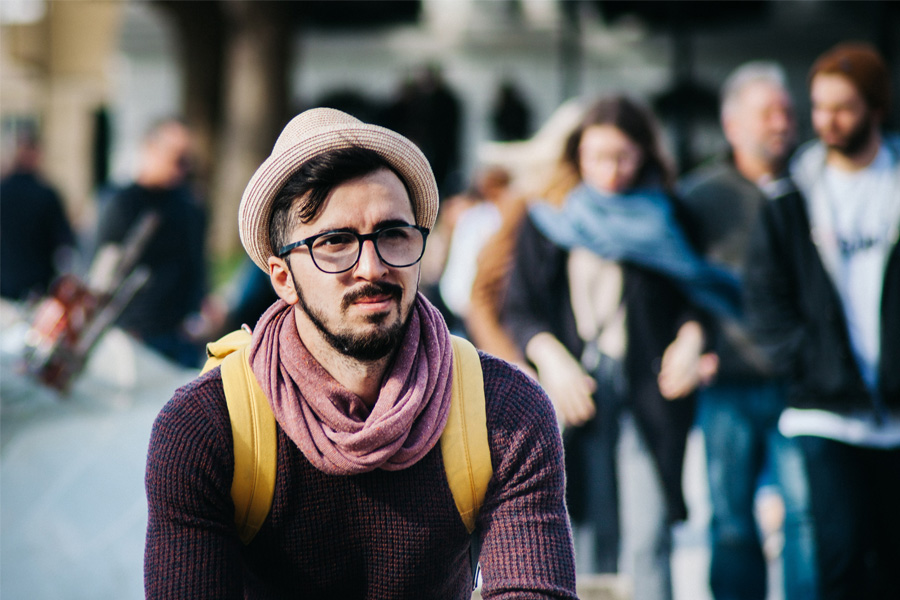 Young man wearing a hat, scarf, glasses and backpack, sits in a public space watching people pass by.