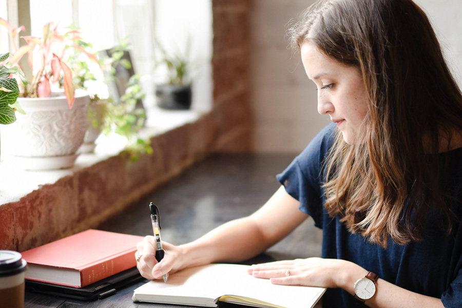 Young woman sits by a window writing in a journal.