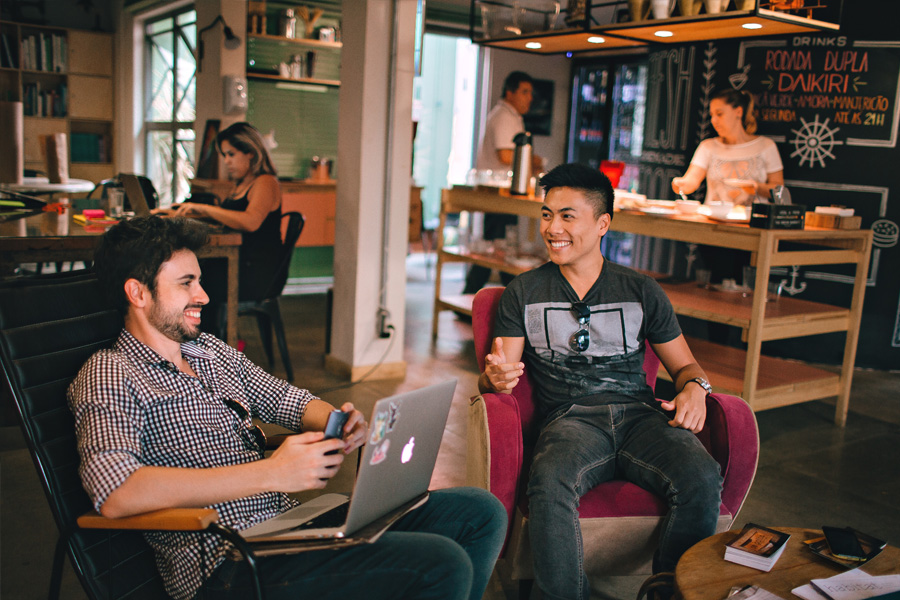 Two excited, young men are having a conversation. One holds a laptop and phone. 