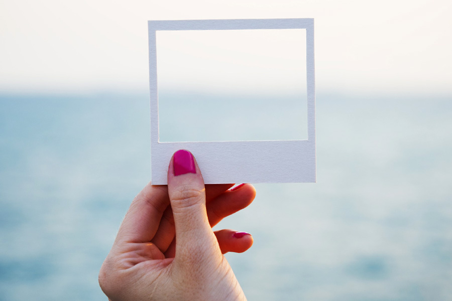 Woman's hand holds a small cardboard frame up to a view of the ocean and uses it to frame part of the horizon.