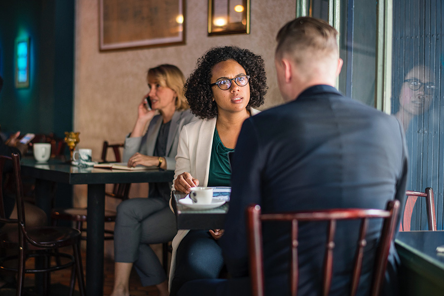 Man and woman in business clothes sit at a café in conversation. We can't see the man's expression however the woman appears to be concerned. 