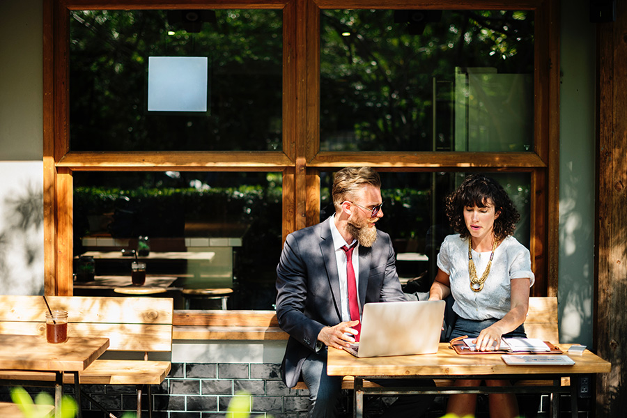 Man and woman in business clothes sit talking at a café. The woman refers to a notebook and the man is using a laptop.