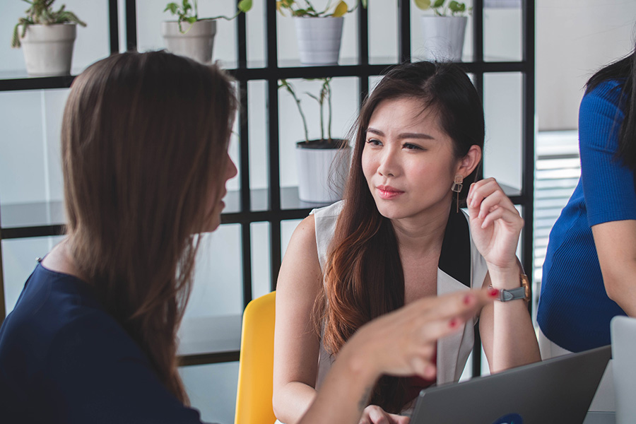 Two women sit together talking in a work or study environment.