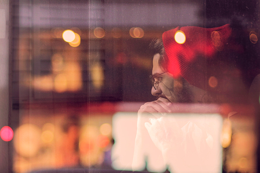 Man sits thinking behind glass reflecting lights and evening street scene.