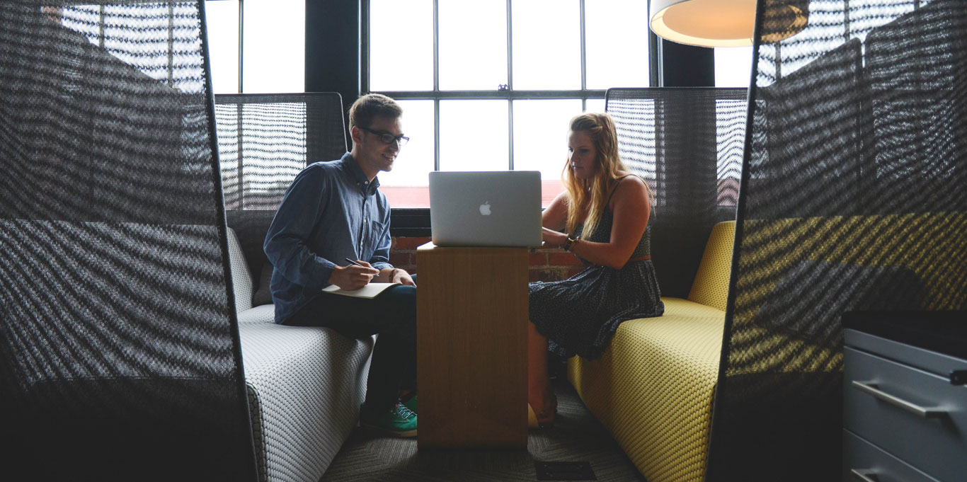 A male and female talking together in a collaborative workspace, an open laptop on the table.