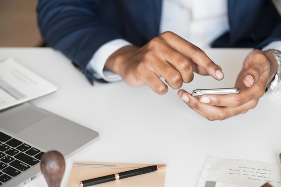 Businessman using a smart phone at a desk with a laptop, note paper and pen.